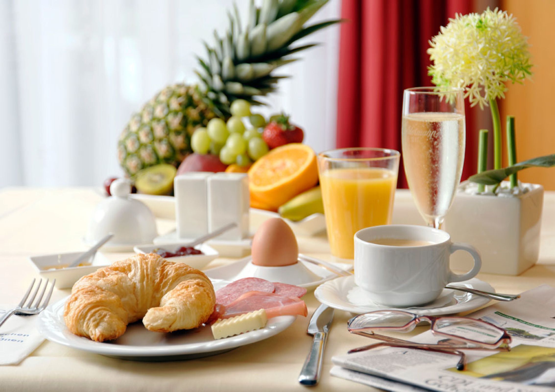 Un desayuno con croissants, huevos, jamón, frutas, jugo y una copa de champán.