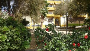 Un edificio amarillo con balcón, rodeado de arbustos verdes y flores rojas.