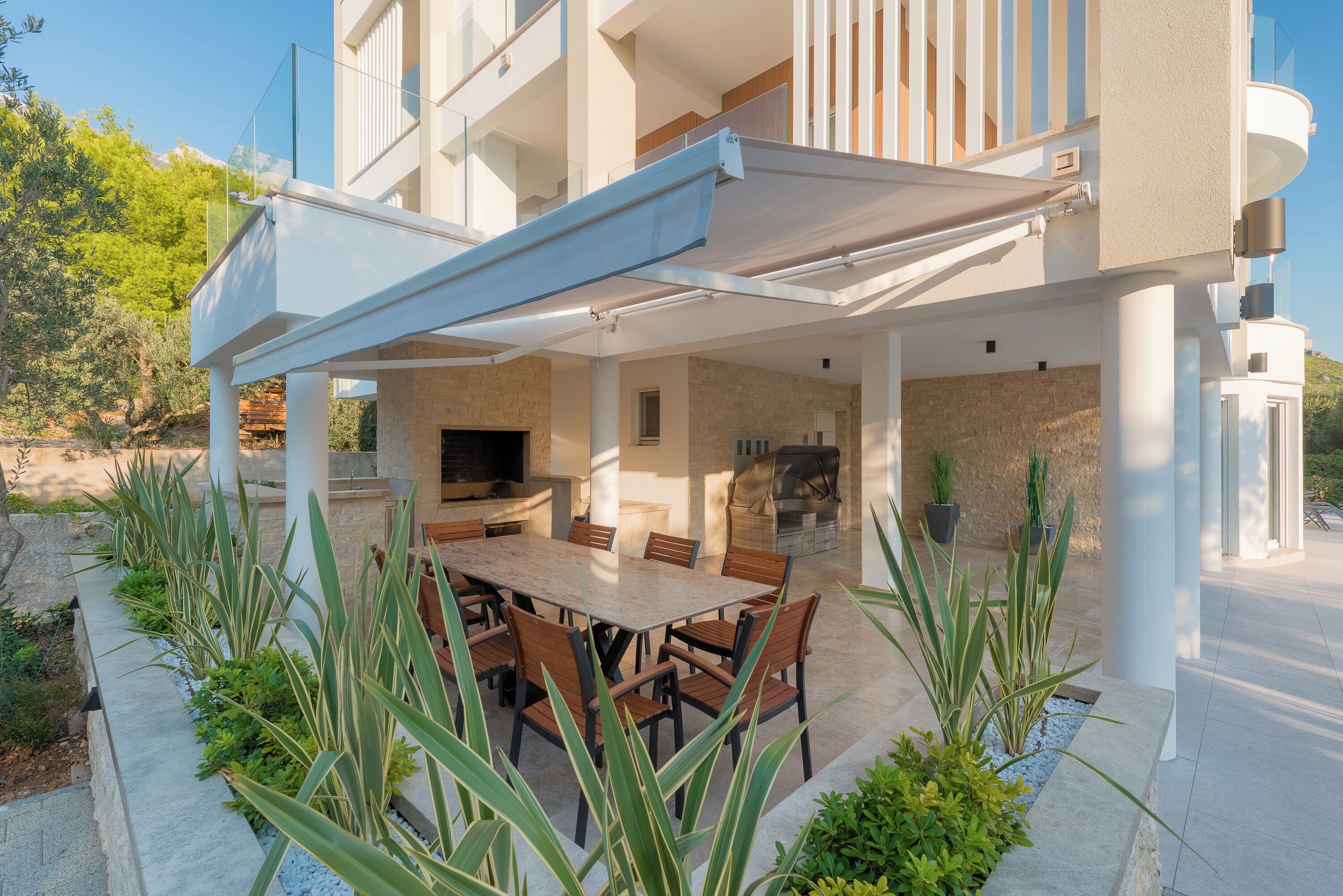 A covered outdoor dining area with a table and chairs, a grill, and potted plants.