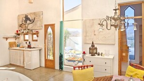 Habitació amb zona de cuina, taula de dinar amb cadires, finestra gran i xemeneia.