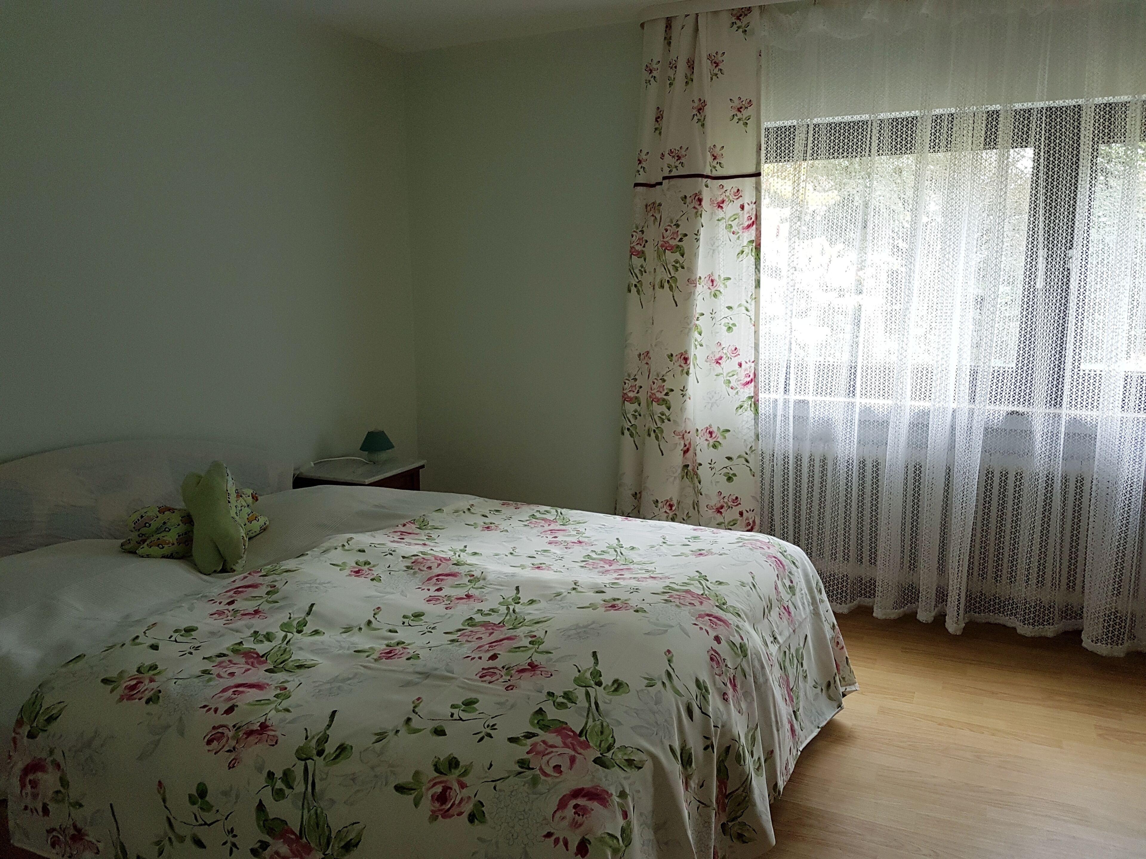 Ein Schlafzimmer mit einer blumigen Tagesdecke, einem Nachttisch mit Lampe und einem Fenster mit durchscheinenden Vorhängen.