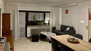 A compact living space with a dining area, a sofa, a radiator, and a window with curtains. - Ferienwohnung Peters – 60m² apartment with WiFi, terrace & nature setting (Schotten)