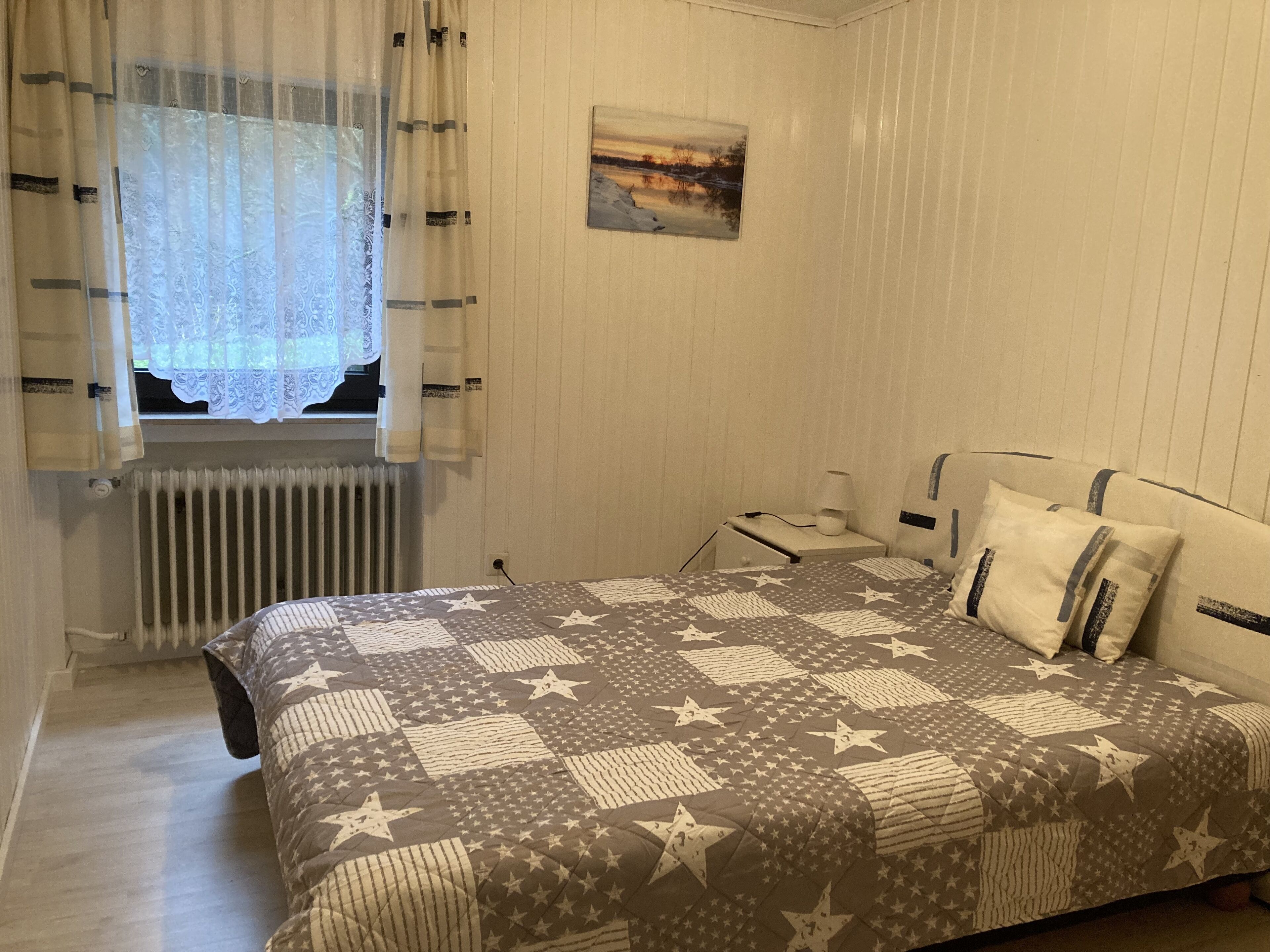 A bedroom with a bed, a nightstand, a lamp, a window with curtains, a radiator, and a painting on the wall.