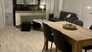 A modern living room with a wooden dining table, black chairs, a grey sofa, and a kitchen area with a radiator and potted plants. - Ferienwohnung Peters – 60m² apartment with WiFi, terrace & nature setting (Schotten)