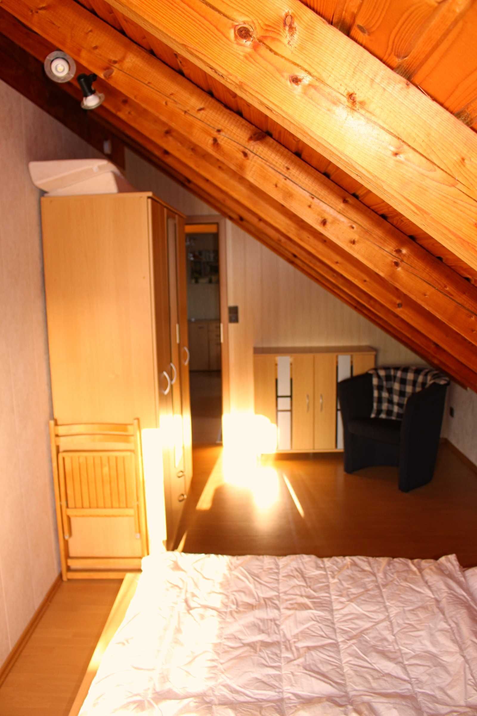 A bedroom with a sloped ceiling, wooden wardrobe, a bed, and a chair.