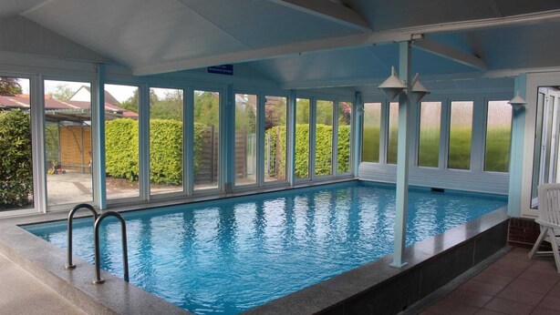 Piscina interior com paredes de vidro transparente, chão revestido de azulejos e uma cadeira branca próxima à piscina.