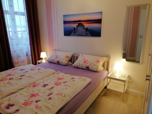 A hotel room with a bed, a nightstand, a lamp, a painting of a sunset, and a mirror.