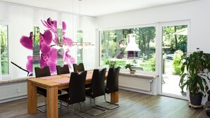 A modern dining area with a wooden table and black chairs, large windows offering a view of the outdoors, and potted plants. - Vacation apartment at Höpen - Lüneburg Heath (Schneverdingen)