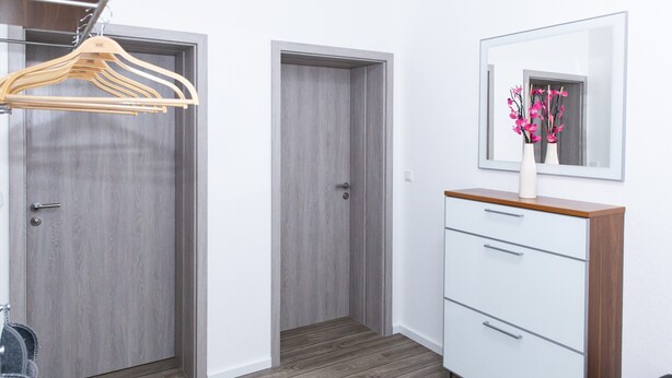 A room with a wooden floor, a white dresser with drawers, a mirror, and a hanging wooden hanger. - Vacation apartment at Höpen - Lüneburg Heath (Schneverdingen)