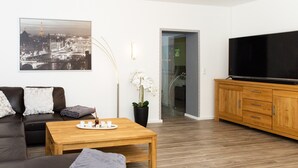 A modern living room with a sofa, a coffee table, a TV, and a painting of a cityscape. - Vacation apartment at Höpen - Lüneburg Heath (Schneverdingen)