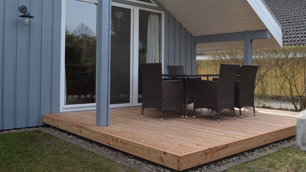 Un porche cubierto con un suelo de madera, una mesa con sillas y una puerta corrediza de vidrio que da a una habitación.