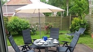Une terrasse avec une table ronde, quatre chaises, une bouilloire bleue et un grand parasol.