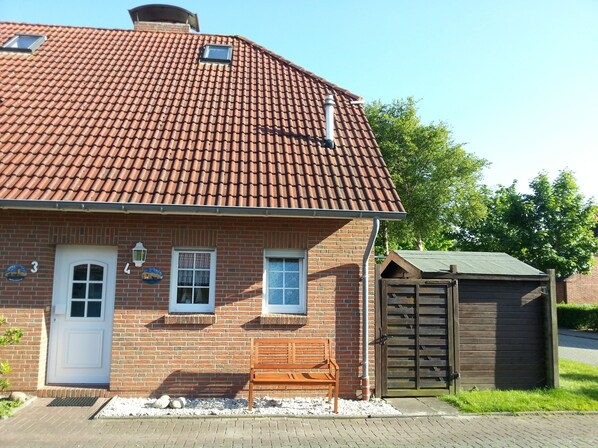 Une maison en briques avec un toit recouvert de tuiles rouges, une porte blanche et un banc en bois à l’extérieur.