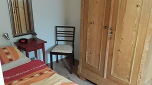 Een slaapkamer met een bed, een houten garderobe, een stoel, een klein tafeltje met een klok en een spiegel aan de muur.