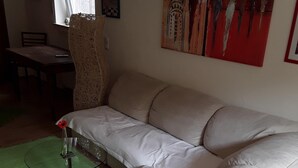 Een kamer met een beige bank, een groen kleed, een glazen tafel met een rode bloevas en een schilderij aan de muur.