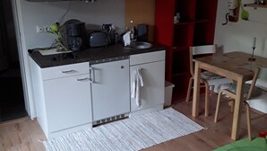 Een compacte keuken met witte kastjes, een magnetron en een koffiezetapparaat. Er is een eethoek met een tafel en stoelen, en een klein rode plank.