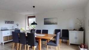 A dining area with a wooden table and chairs, a black pendant light, a window with blinds, a painting on the wall, and a kitchenette with cabinets and a countertop. - Large, cozy vacation home with garden between St. Peter-Ording & Büsum (Wesselburen)