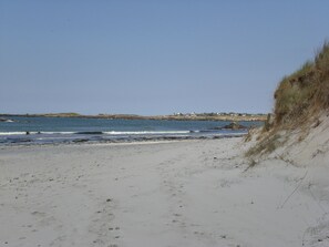 Une plage de sable avec des vagues, des dunes herbeuses et une côte lointaine parsemée de bâtiments.