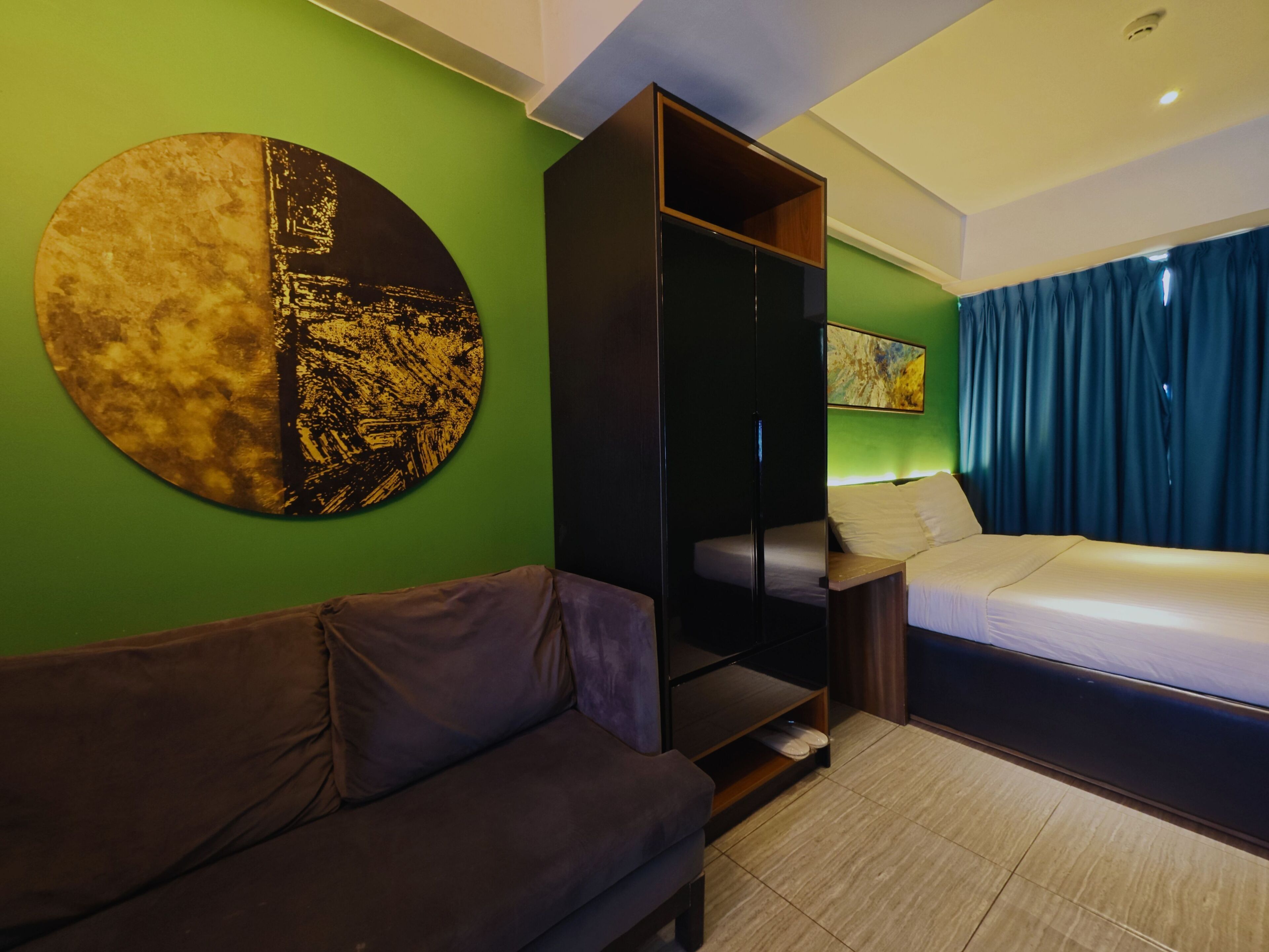 A modern hotel room with a green wall, a round artwork, a sofa, a bed, and a wardrobe.