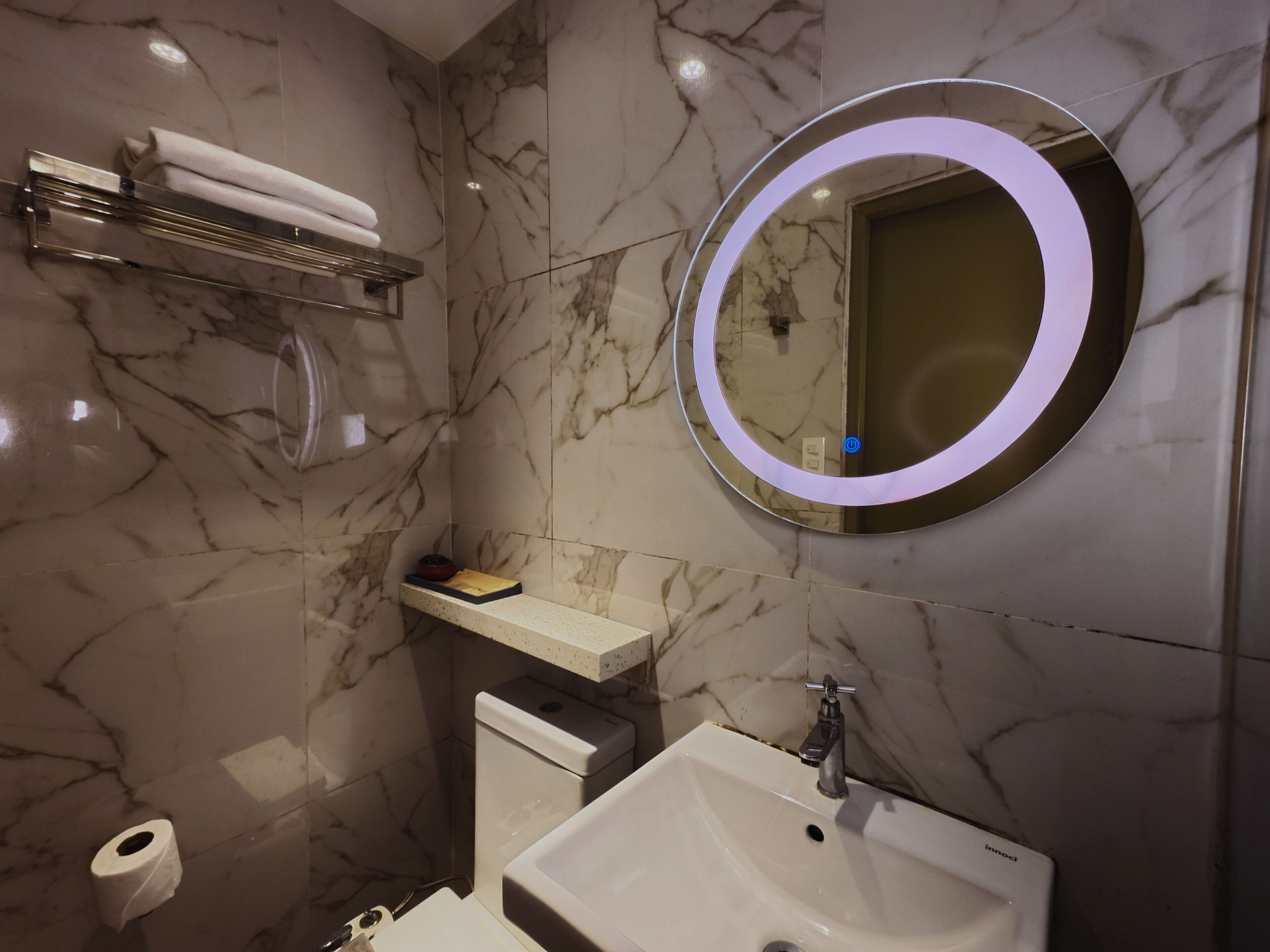 Um banheiro com parede de mármore, pia, vaso sanitário, porta-toalhas e espelho iluminado.