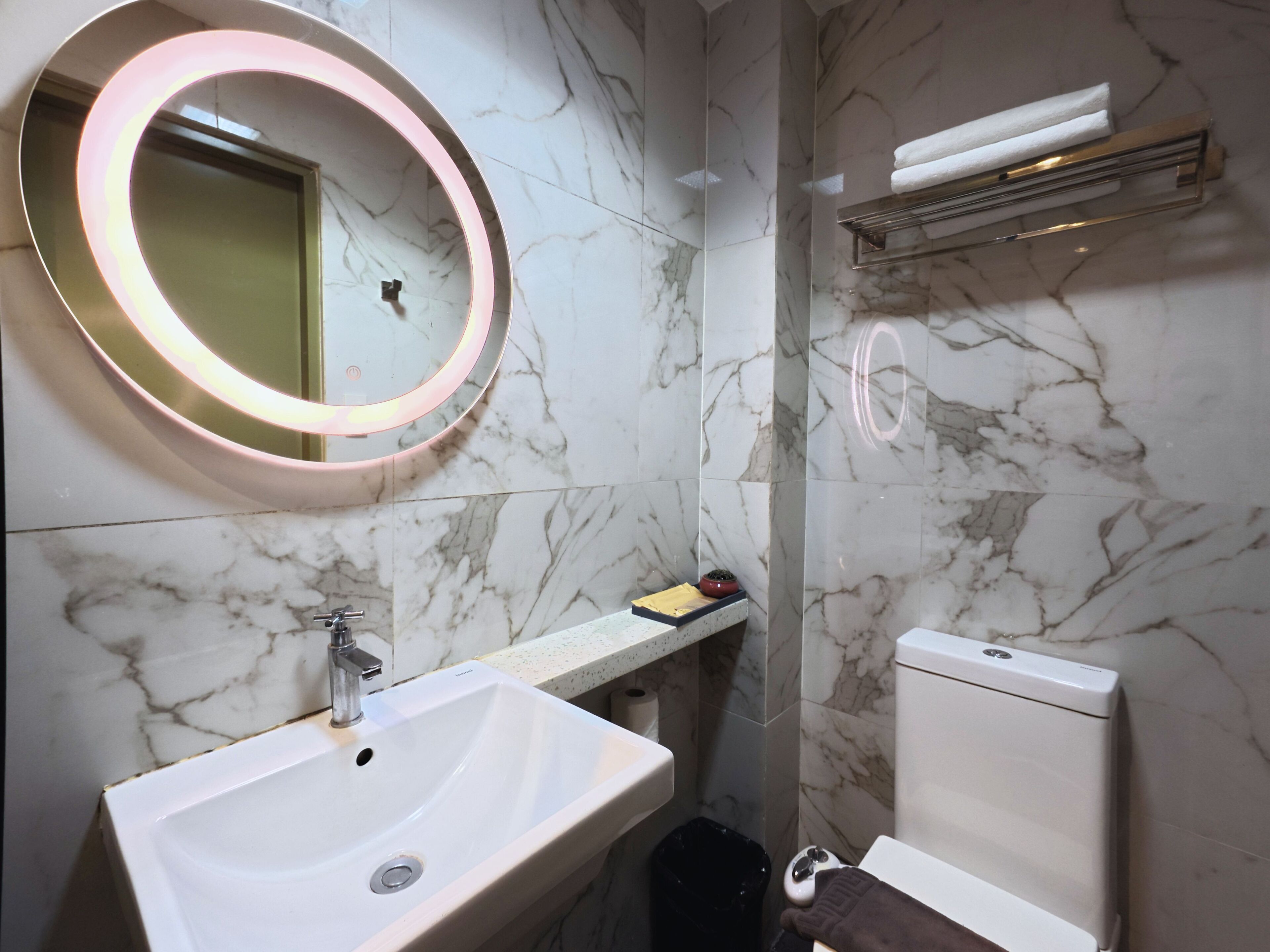 A bathroom with a white sink, a round mirror with a light, a toilet, and a trash bin.