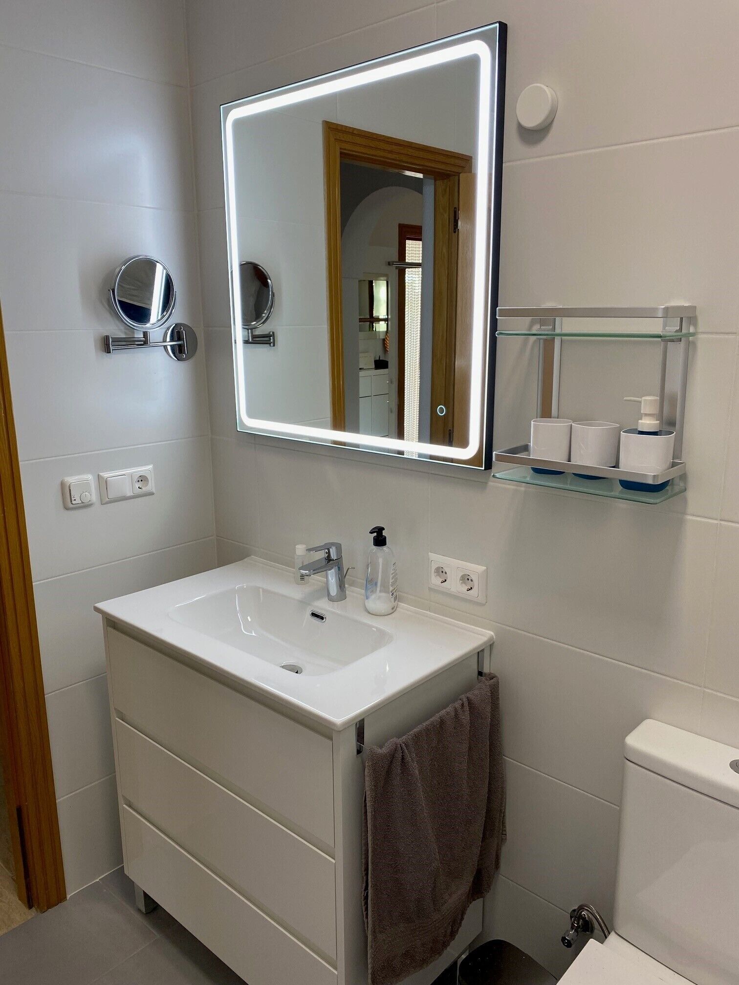 A modern bathroom with a white sink, a mirror, a towel rack, and a glass shelf with toiletries.