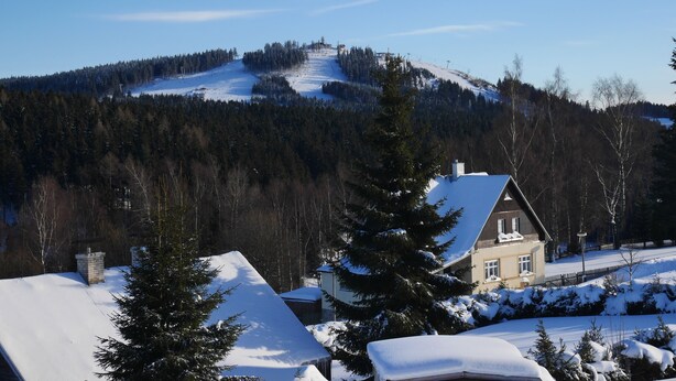 Ein Haus mit geneigtem Dach und Schornstein, umgeben von schneebedeckten Bäumen und einem Berg im Hintergrund.