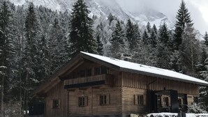 A wooden cabin with a sloped roof, surrounded by snow-covered trees and mountains.