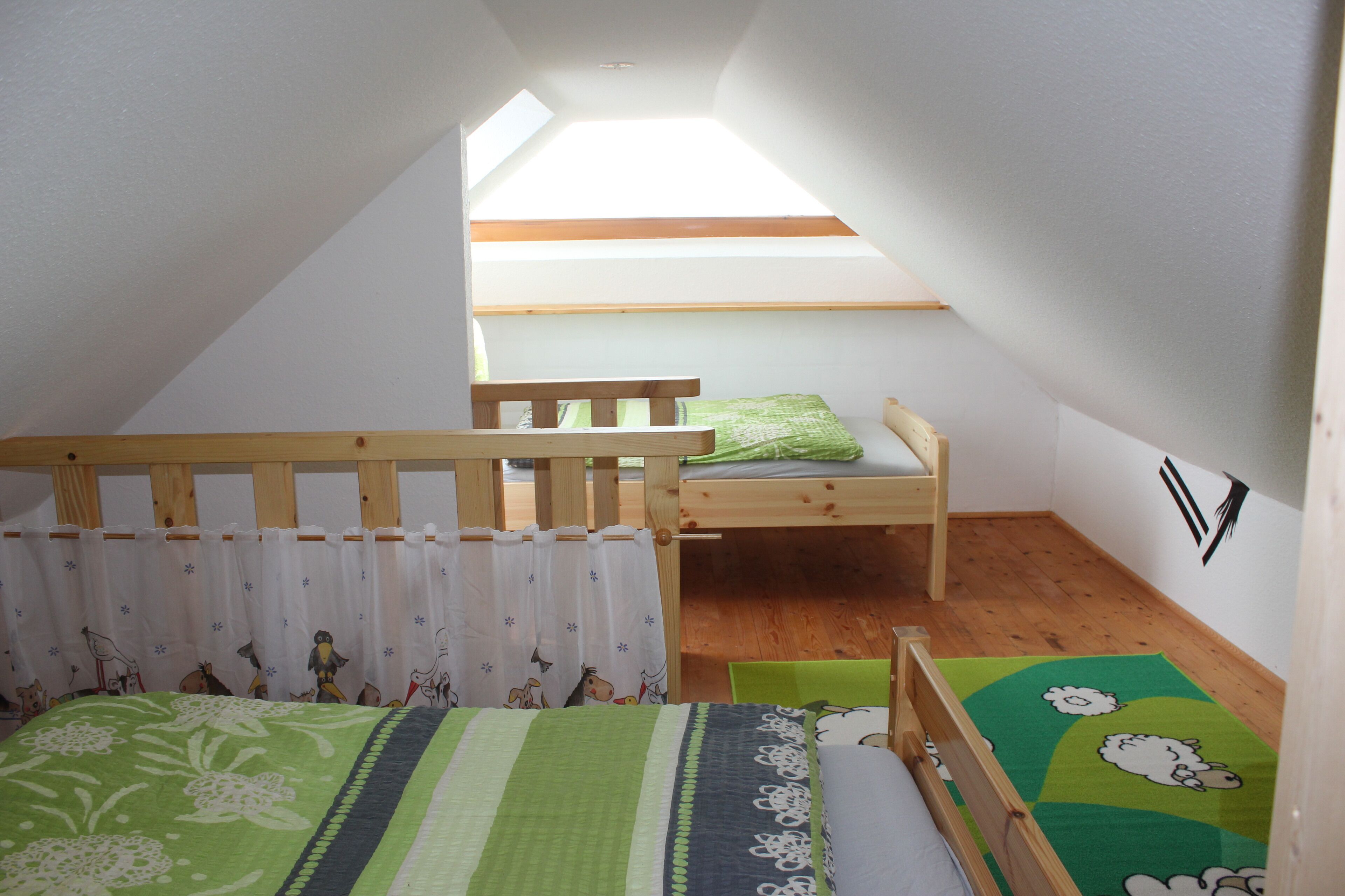 A small, cozy attic bedroom with a sloped ceiling, a wooden bunk bed, a single bed with a green and white floral bedspread, a wooden chair, and a small rug with cartoon characters.