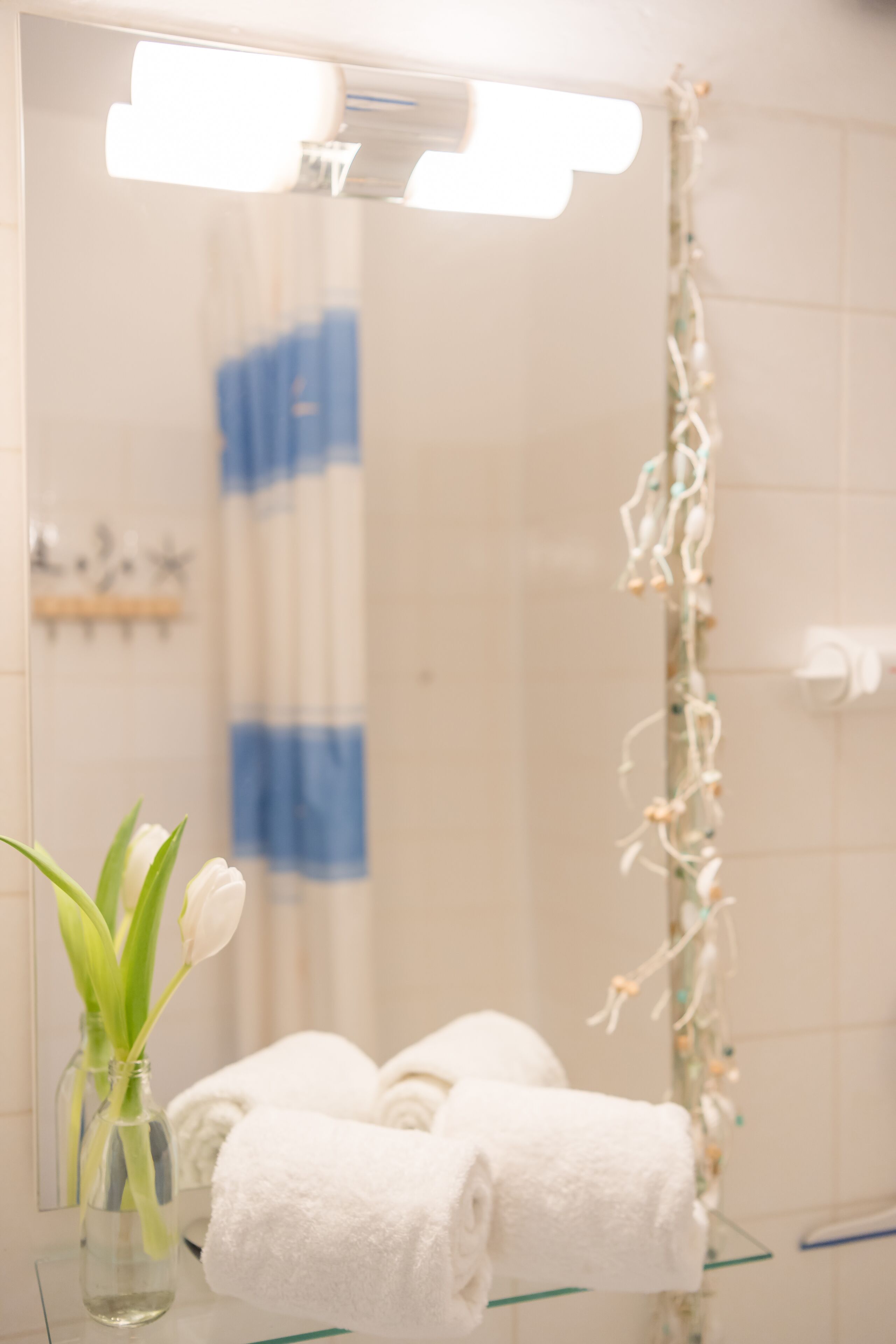 Une salle de bain avec une douche à parois de verre, une serviette blanche posée sur une étagère en verre et un vase contenant une seule fleur blanche.