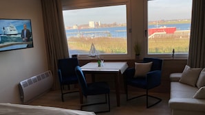 A hotel room with a bed, a desk with a chair, a small table with a bottle, and a view of a body of water and buildings.