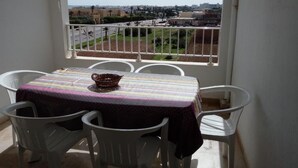 Un balcon avec une table et des chaises, donnant sur une rue bordée d’immeubles et d’espaces verts.