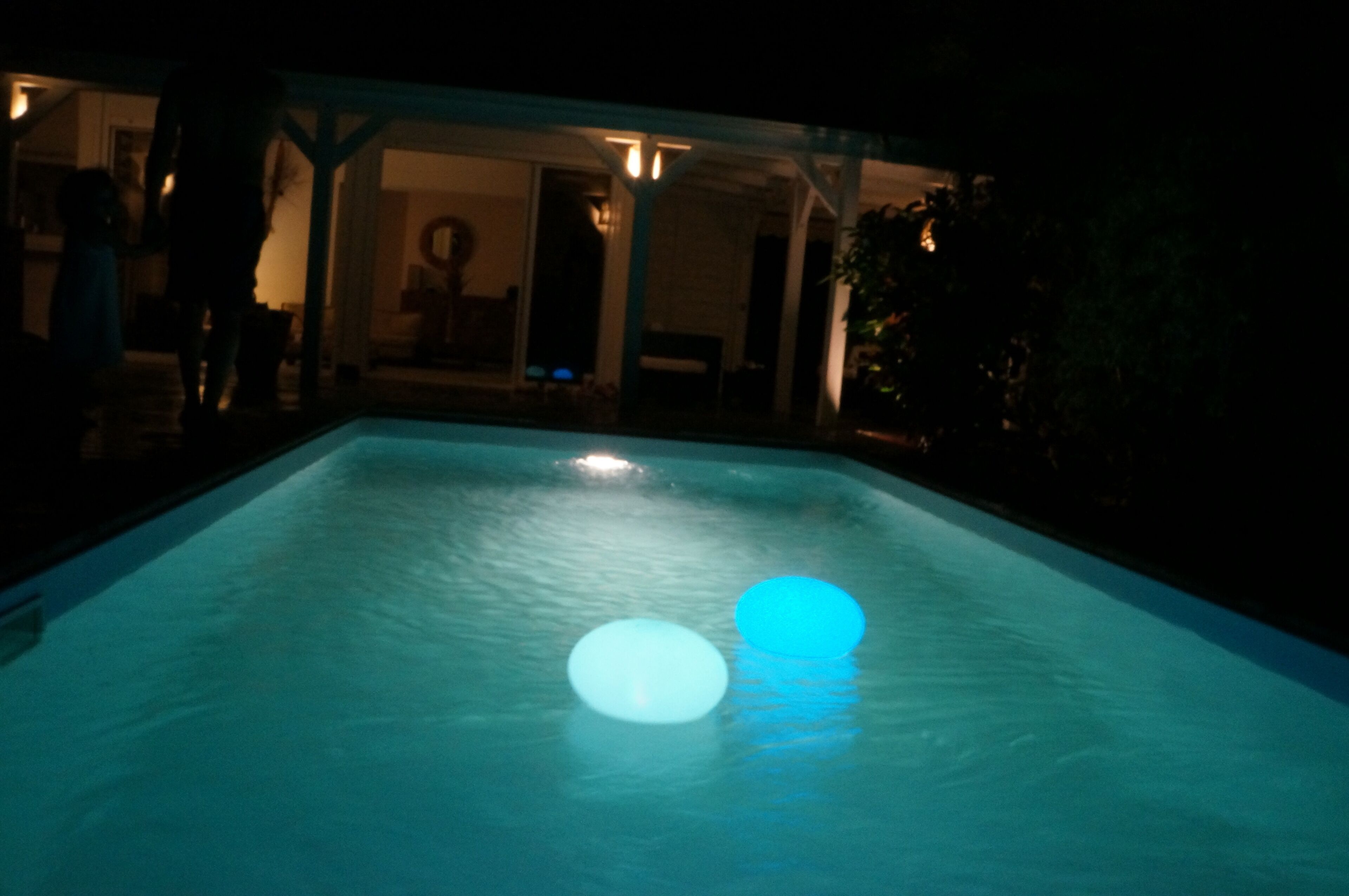 A swimming pool with two illuminated balls, surrounded by a covered area with a view of a building in the background.