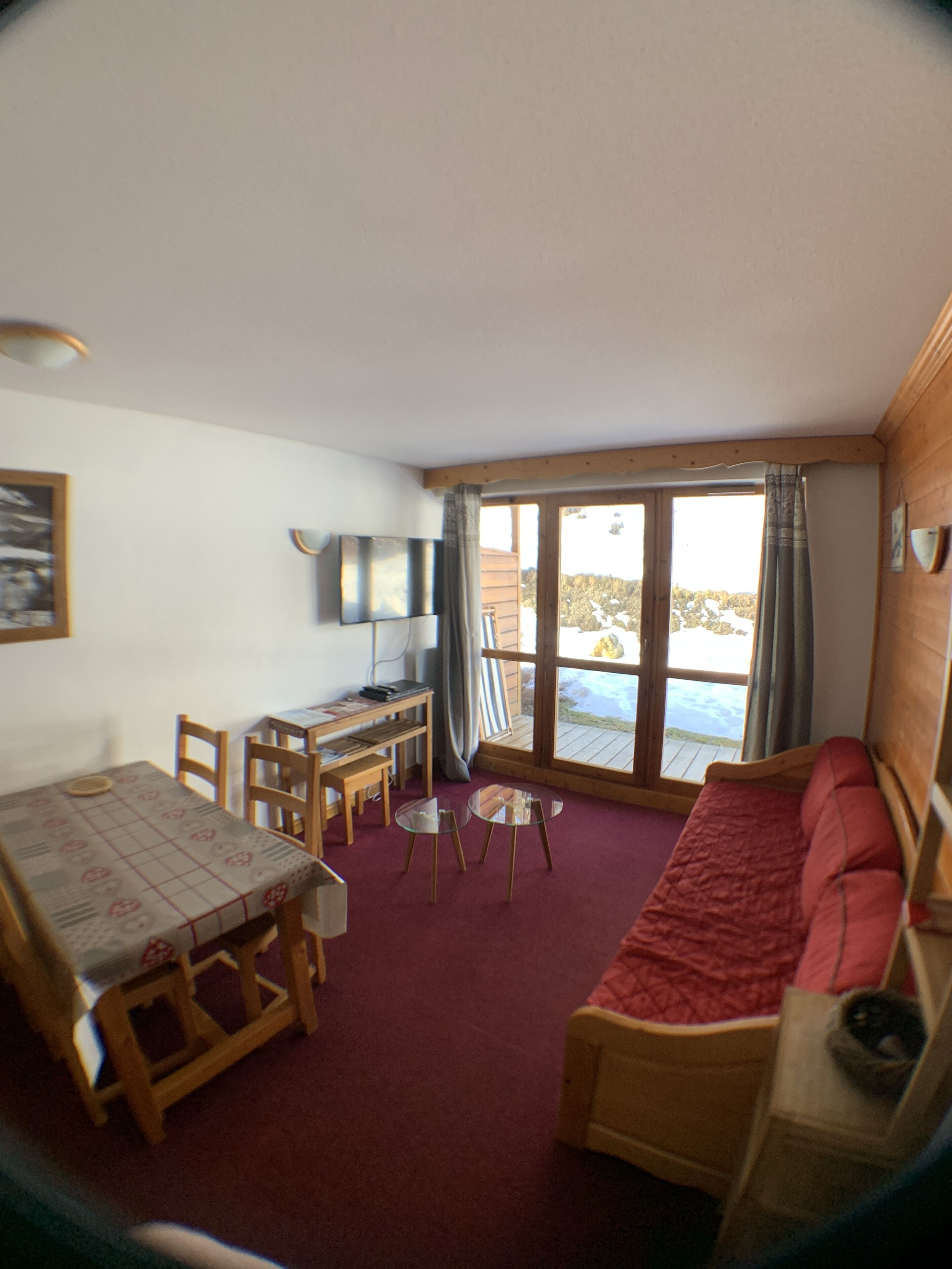 A cozy room with a wooden sofa, a dining table with chairs, a TV, and a large window with curtains.