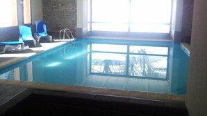 Una piscina interior con paredes de vidrio transparente, sillones reclinables azules y una pared de piedra.