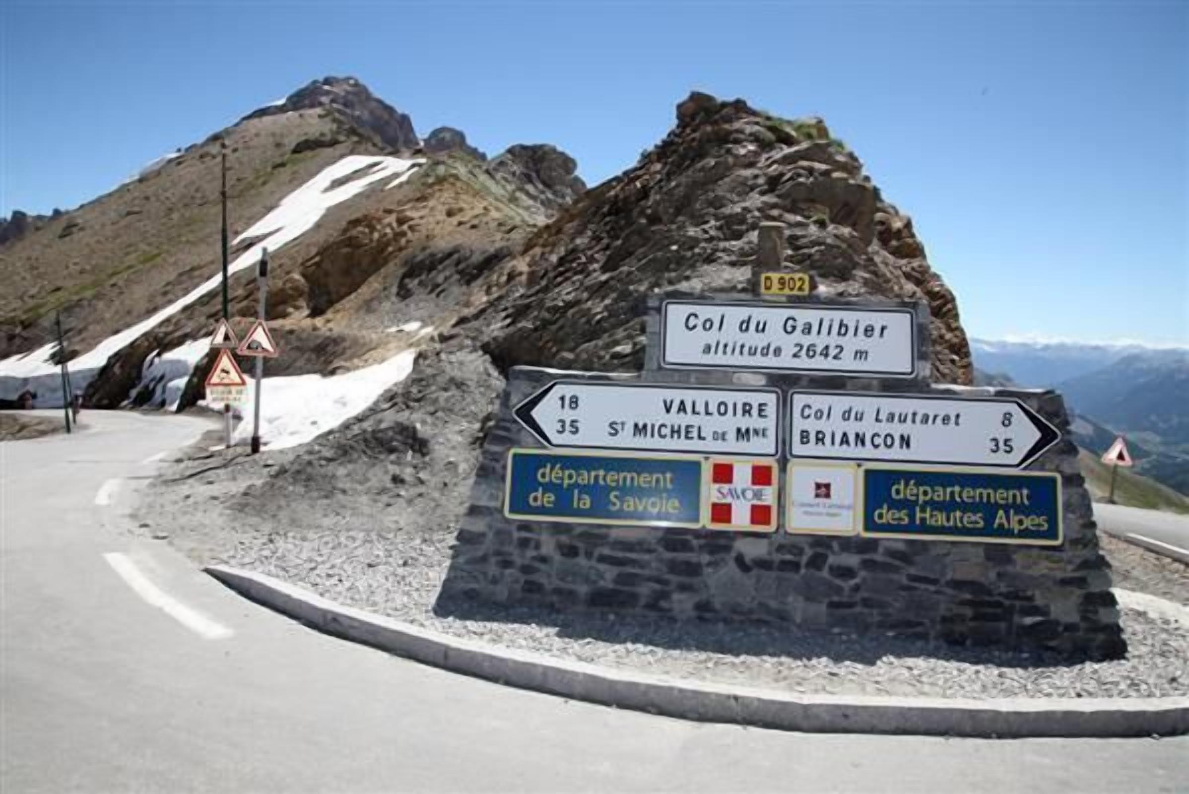Un panneau de signalisation directionnel à un carrefour sur une route de montagne, indiquant plusieurs destinations.