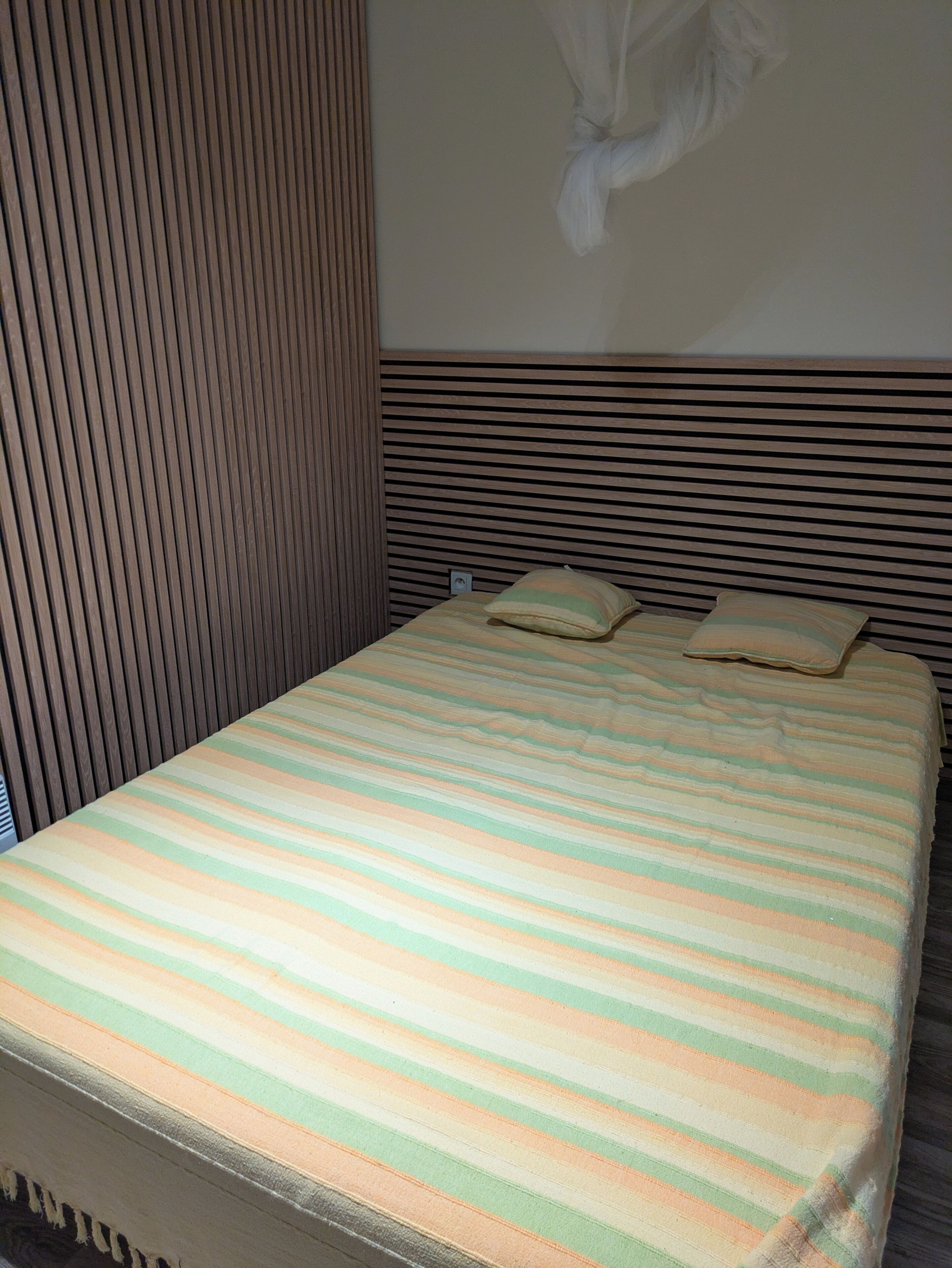 A bed with a striped bedspread, a headboard with horizontal slats, and a wall-mounted light fixture.