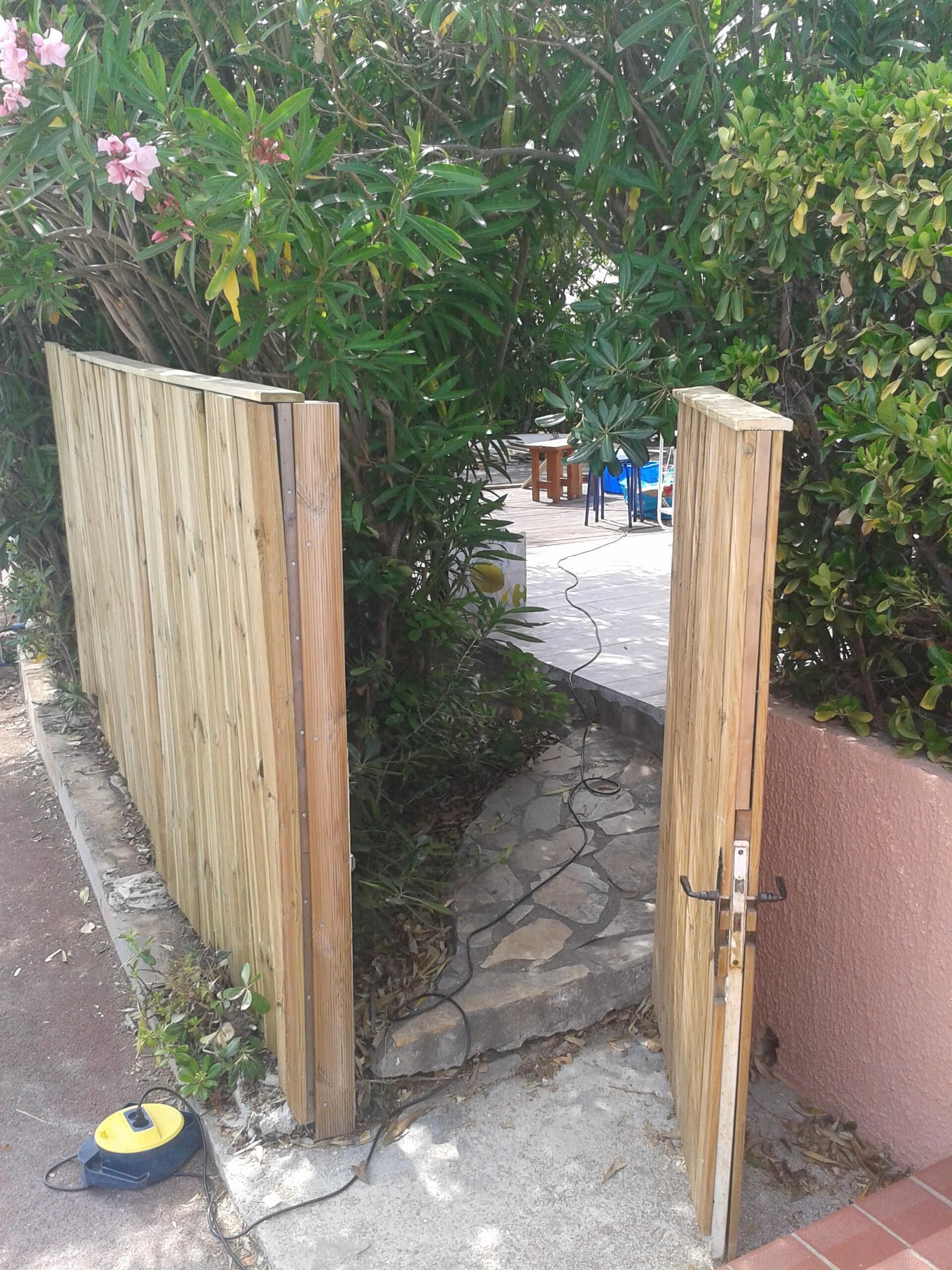 A wooden gate leading to a paved pathway surrounded by greenery and a stone wall.