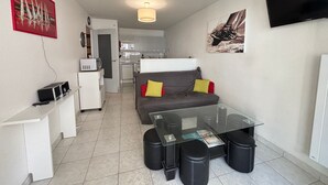 Un espace de vie compact comprenant un coin cuisine, un canapé avec des coussins, une table basse en verre et un téléviseur à écran plat fixé au mur.