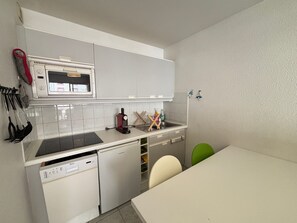 Een kleine keuken met een vaatwasser, een magnetron en een spoelbak. Er hangen keukengerei aan de muur en er staat een groene stoel bij de tafel.