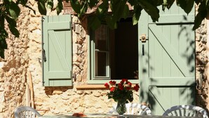 A rustic stone building with a window and shuttered door, a table with a floral tablecloth, and a vase with red flowers. - Le Jas, charming Mas in Provence with shared pool, nature, calm, space (Flayosc)