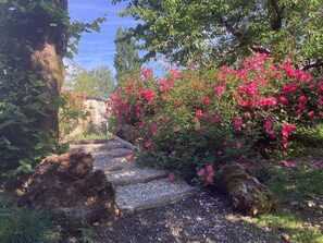A garden pathway with a stone wall, surrounded by lush greenery and pink flowers. - Longère du Marais Poitevin (La Ronde)