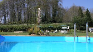 A clear blue swimming pool with steps leading into it, surrounded by greenery and a few outdoor chairs and umbrellas.