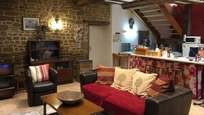 A cozy living room with a stone wall, wooden beams, a sofa with red and beige cushions, a coffee table, and a kitchen area with a microwave and a wooden counter.