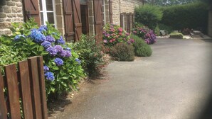 A stone building with wooden shutters, a paved driveway, and a variety of flowering plants.