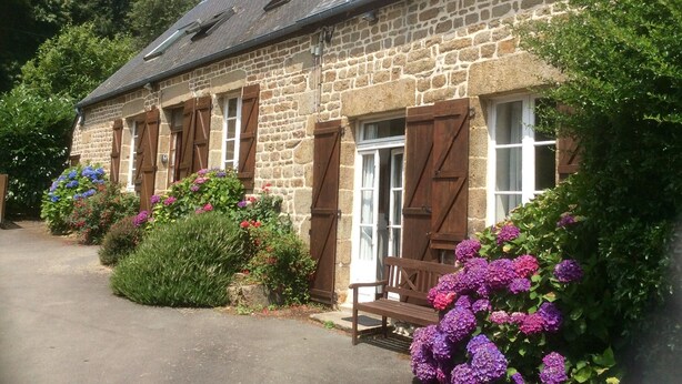 A stone building with wooden shutters, a bench, and flowering plants.