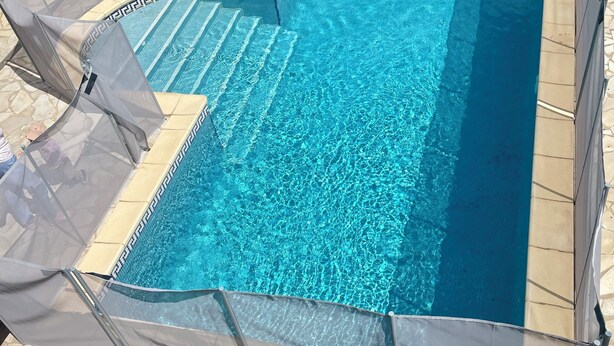 A rectangular swimming pool with clear blue water, surrounded by a stone deck and a white fence.