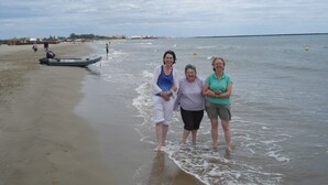 A beach scene with three people standing in shallow water, a boat on the sand, and a cloudy sky.