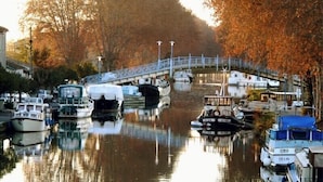 A canal with boats docked, a bridge, and trees with autumn leaves.