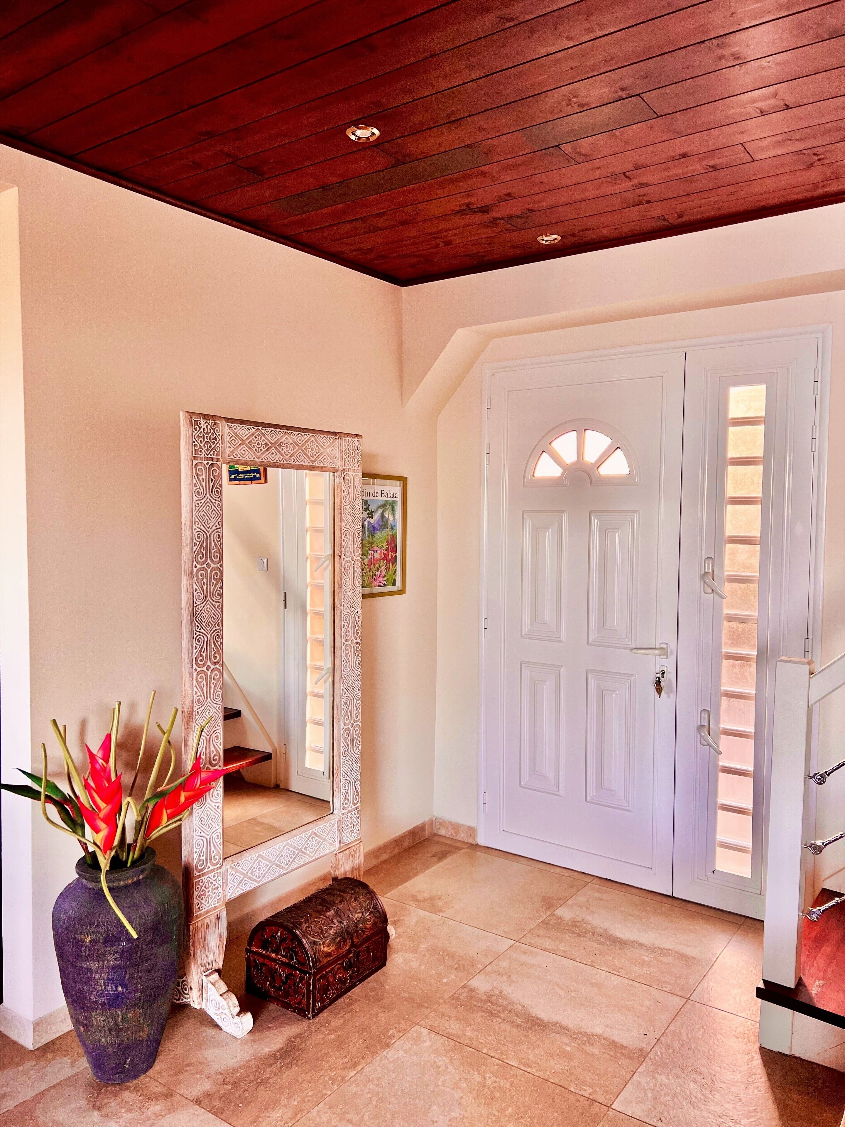 Un corridoio con soffitto in legno, una porta bianca con arco decorativo, uno specchio grande con cornice a motivi e un vaso con fiori rossi.